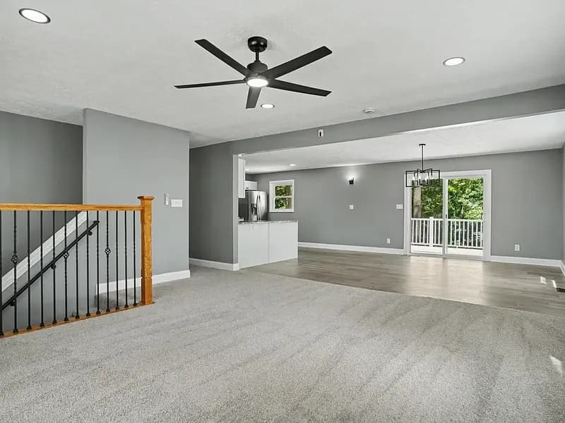 Carpeted living room with a black ceiling fan and a stair railing with a wood handrail and black balusters, opening to a dining area with a sliding door to a deck