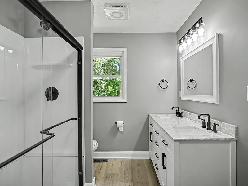 Bathroom with a double-sink white vanity, marble-look top and black faucets, a white framed mirror, a window, and a glass shower with a black frame