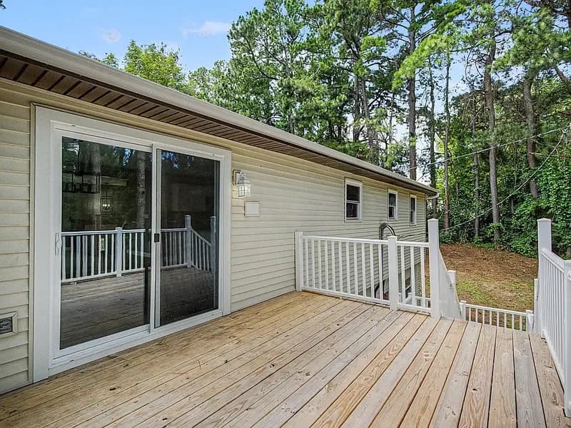 Wood deck with white railings beside a sliding glass door on light siding, with tall pine trees beyond