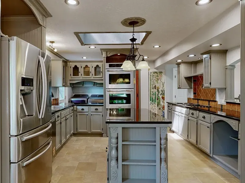 Kitchen with gray painted cabinets, black countertops, a stainless French-door refrigerator, double wall ovens, and a center island with open shelves and turned posts