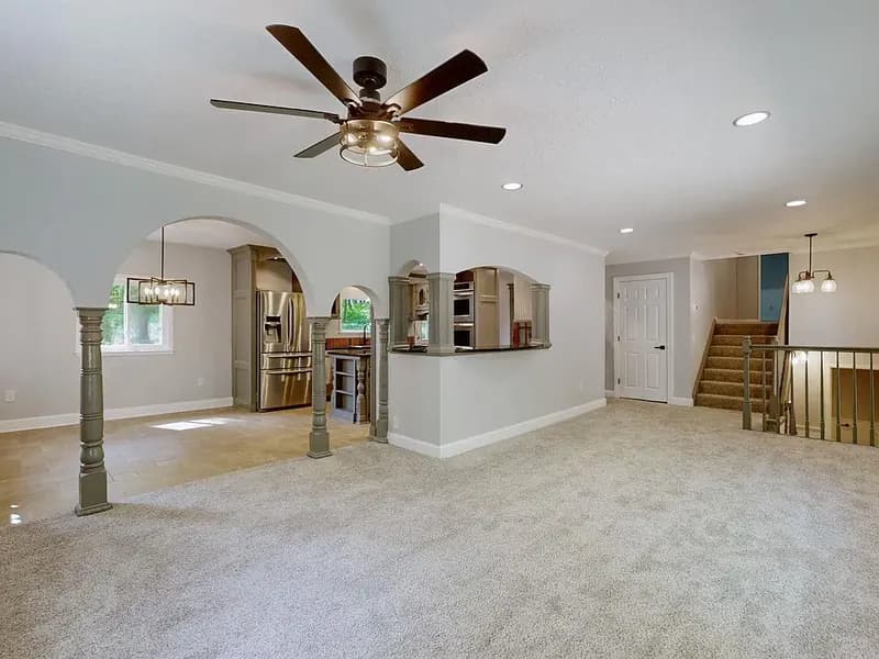 Carpeted great room with a ceiling fan and arched openings on columns into the dining area and kitchen, with a stairway and railing at the right