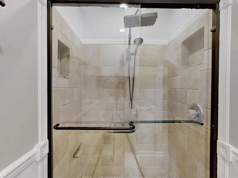 Shower with beige tile walls, two recessed niches, a rain showerhead and a handheld sprayer, behind dark-framed sliding glass doors