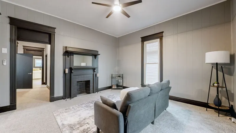 Living room with a black fireplace surround and mantel mirror, light gray paneled walls with dark trim, carpet, a ceiling fan, and a gray sofa, rug and floor lamp