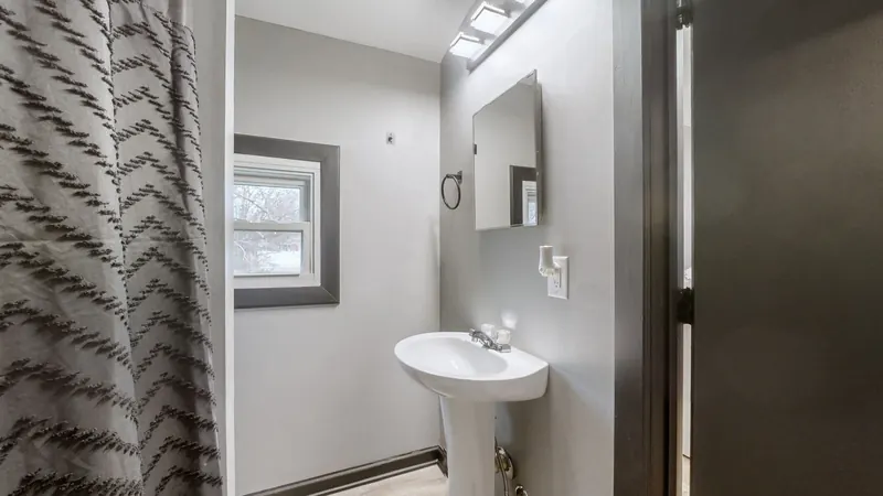 Bathroom with a white pedestal sink, a frameless mirror, a vanity light bar, a window with dark trim, and a patterned gray shower curtain