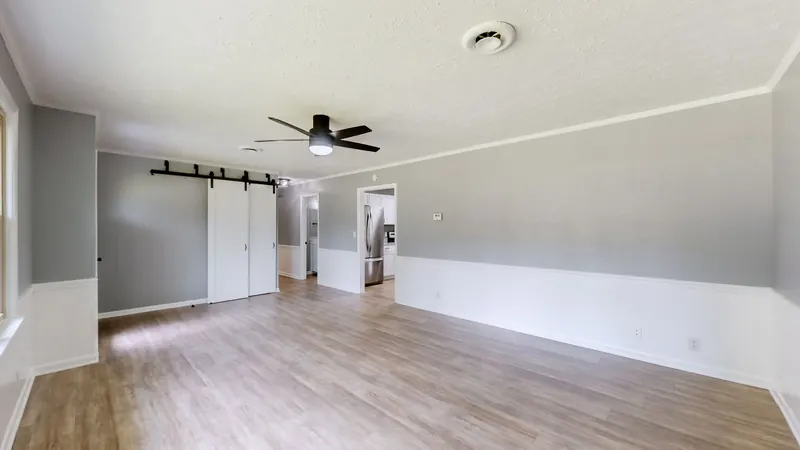Empty living room with gray walls over white wainscoting, wood-look plank floors, a black ceiling fan, and white sliding barn doors on a black rail