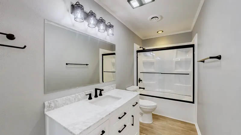 Bathroom with a white vanity and marble-look top, black faucet and hardware, a large mirror under a four-light fixture, a toilet, and a tub-shower with sliding glass doors