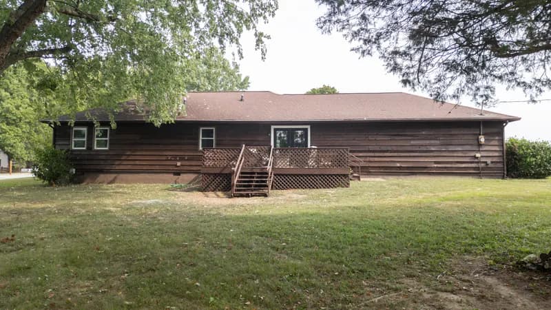 Back of a one-story house with dark horizontal siding, a wood deck with lattice skirting and steps up to glass double doors, and a wide lawn under shade trees