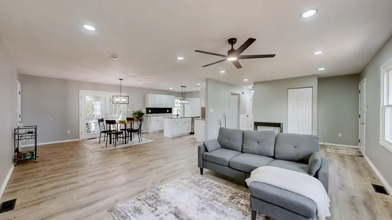 Open living room with light wood-look floors, a gray sofa with a white throw, a ceiling fan and recessed lights, looking toward a dining table and a white kitchen