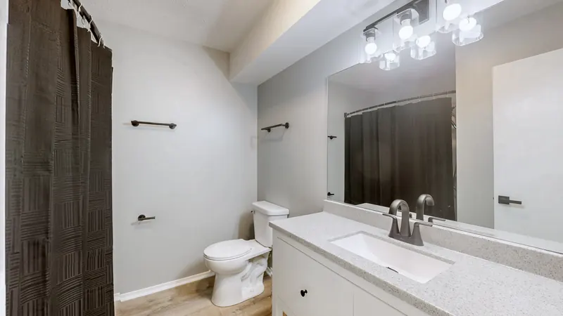 Bathroom with a white vanity and speckled countertop, a dark faucet, a wide mirror under a glass-shade light bar, a toilet, and a dark shower curtain